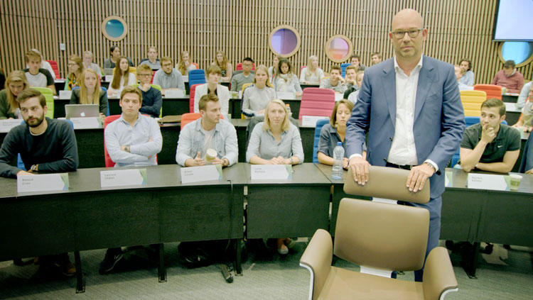 Person stands behind his desk chair in an auditorium with students.