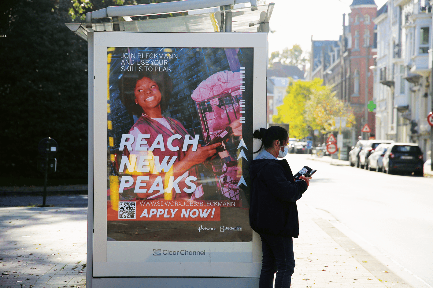 Straataffiche 'Reach New Peaks – Apply Now!' op een Clear Channel abri in een Belgische stad